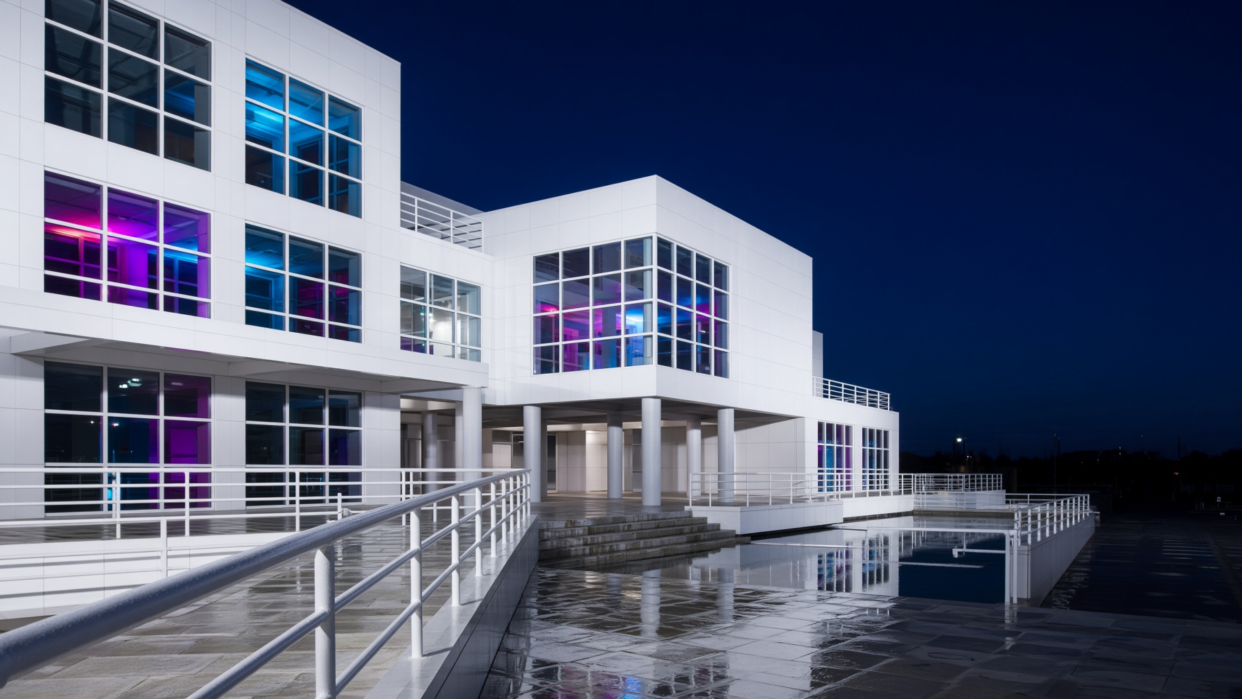 Night view along white pipe railings toward square windows glowing magenta and cyan.